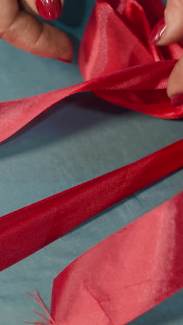 hands of female person tying up bow from red ribbon on blue present box. woman prepares secret gift for birthday celebration of daughter closeup