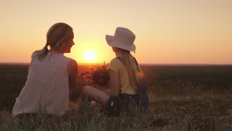 mom and little daughter are sitting on a hill admiring the sunset in the hands of wild flowers summe