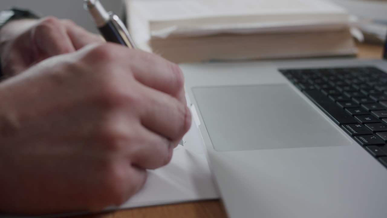 We zien handen die op papier schrijven, terwijl een andere hand een laptop gebruikt. Dit gebeurt in een lichte werkruimte. De activiteit vindt plaats tijdens de werkuren