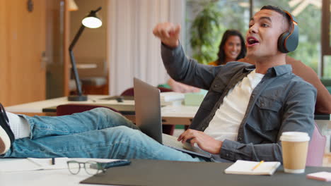 young businessman with feet on desk in office wearing wireless headphones  greeting colleague