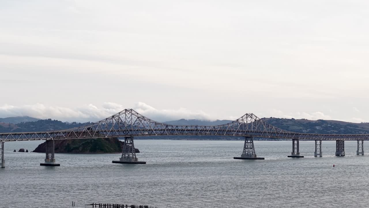 Real time aerial view of the Richmond–San Rafael Bridge stretching across the bay, linking two cities with calm waves below and distant hills bathed in warm light.