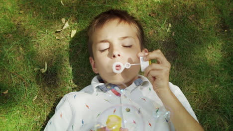 Cheerful-boy-lying-on-green-grass.-Adorable-child-blowing-soap-bubbles-at-camera