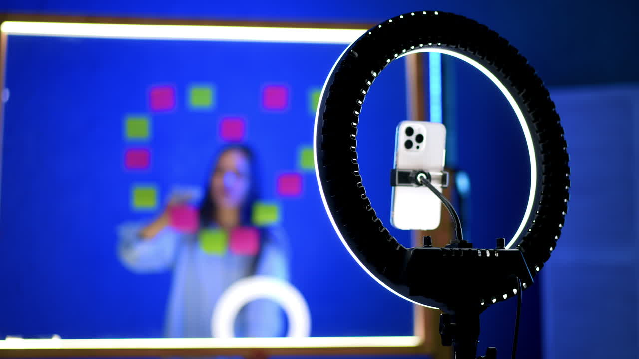 Tripod with a phone and light ring on. Woman stands behind the glass board with stickers on at blurred backdrop.