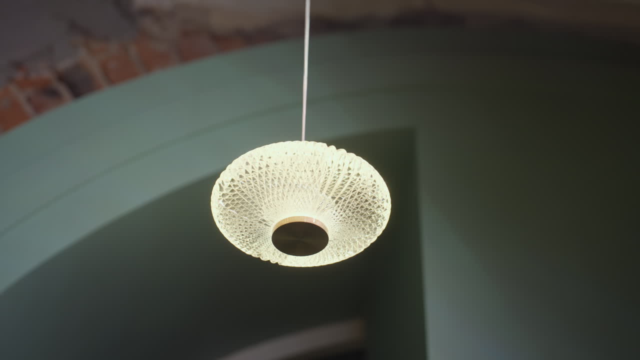 Modern pendant light with intricate glass texture glows warmly while suspended from ceiling inside stylish space featuring soft green tones and exposed brick accents adding charm to ambient setting