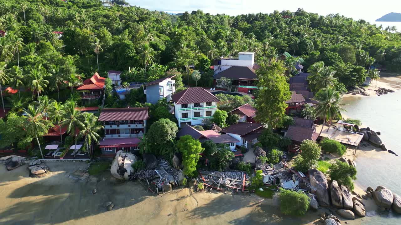 Aerial view of a coastal area with buildings surrounded by lush vegetation and a beach, featuring rocks and calm water.