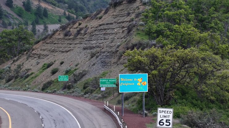 Segnali di Benvenuti in California e Purple Heart Trail con limite di velocità 65 lungo un'autostrada tortuosa, pendio roccioso a strati, alberi verdi sotto un cielo nuvoloso e coperto. California, USA. Vista aerea