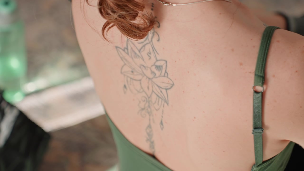Close-up de tatuagem nas costas de mulher em treino em casa, mostrando alça de top esportivo verde e tatuagem floral, gotas de suor na pele, ajuste do ombro durante o desaquecimento pós-treino, com luz natural suave vinda da janela do banheiro.