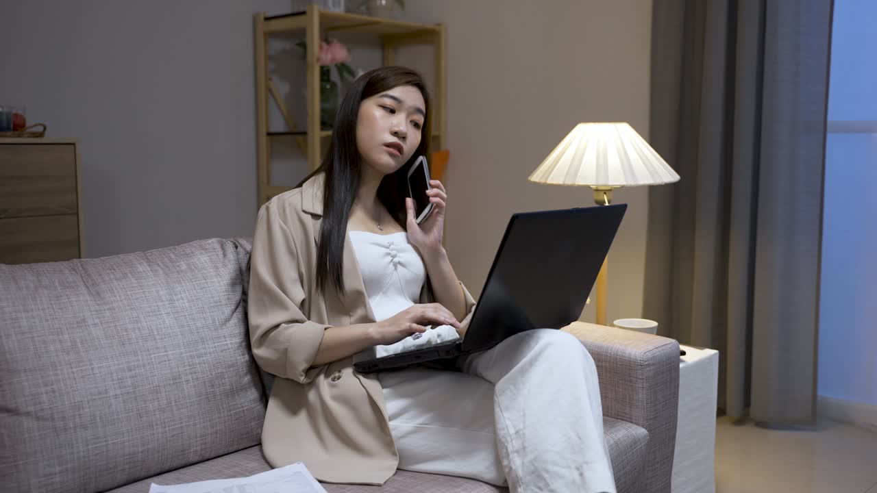 아름다운 아시아 여성 사업가는 집에서 거실의 소파에 노트북에서 오버타임을 하는 동안 전화에 응답하고 있습니다.