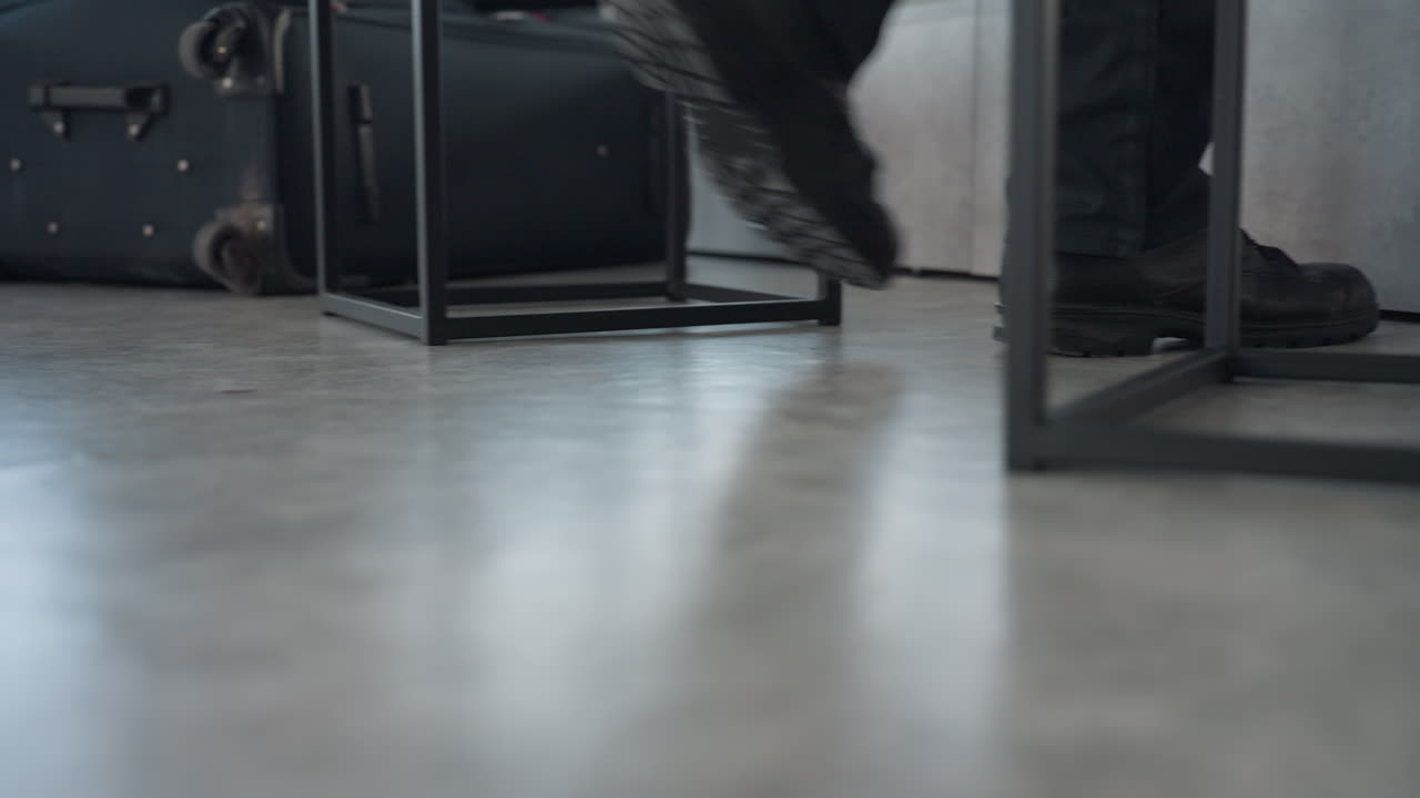 low angle view of person wearing black boots and dark trousers walking across smooth floor toward chair in minimalist room with large window light and mirror reflecting part of scene