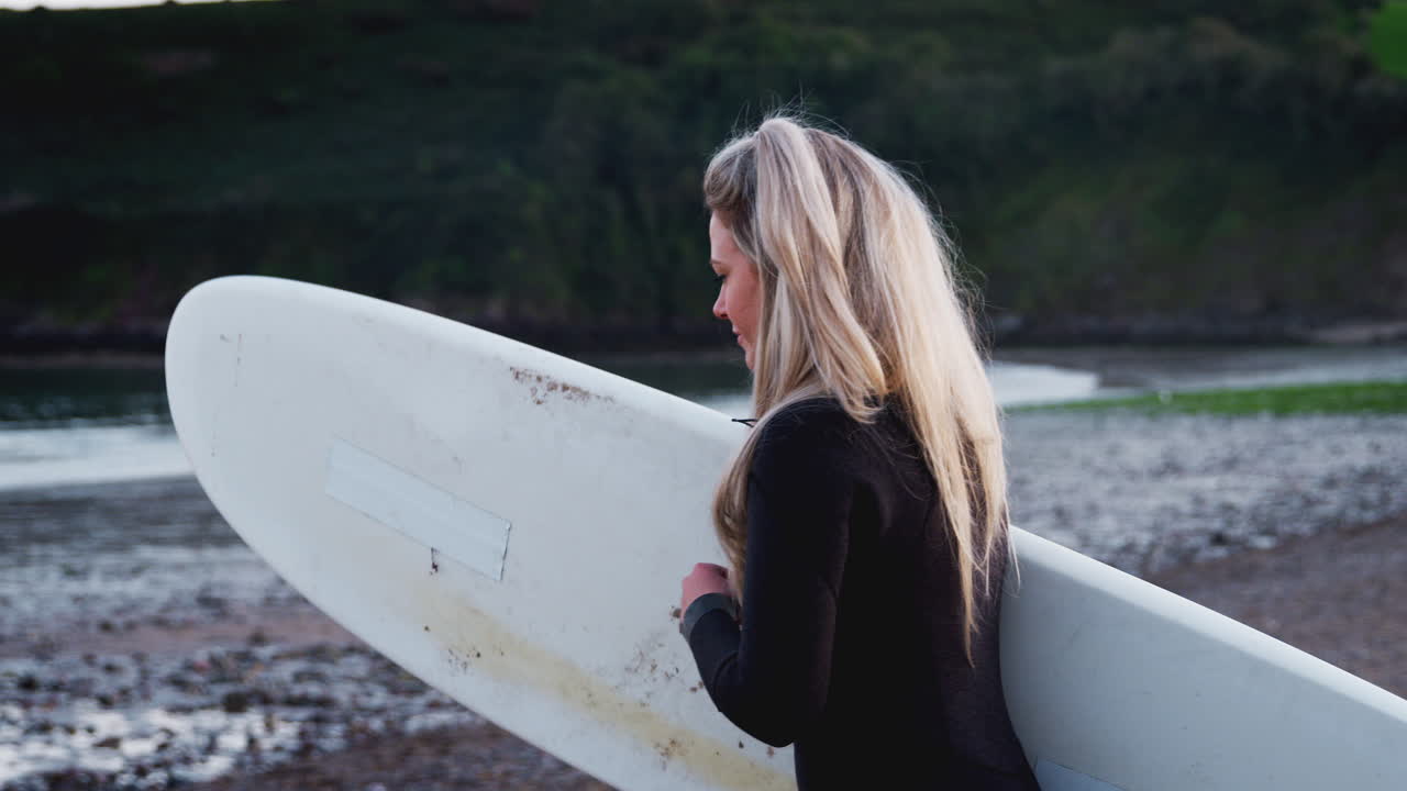 웨이트 슈트를 입은 여성이 해변을 따라 바다로 고 서핑 보드를 들고 서핑 휴가를 즐기고 있습니다.