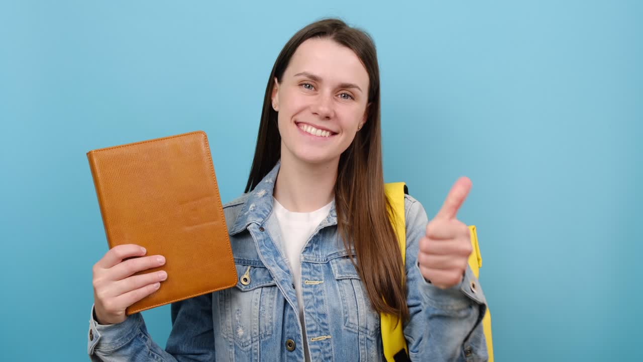retrato de una niña adolescente feliz señalando con el dedo el libro, mostrando el pulgar hacia arriba, lleva una mochila amarilla, posando aislada en un fondo de color azul en el estudio. educación en la escuela secundaria concepto universitario