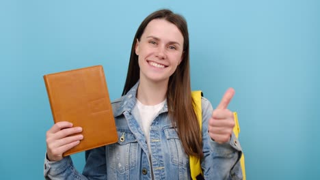 retrato de una niña adolescente feliz señalando con el dedo el libro, mostrando el pulgar hacia arriba, lleva una mochila amarilla, posando aislada en un fondo de color azul en el estudio. educación en la escuela secundaria concepto universitario