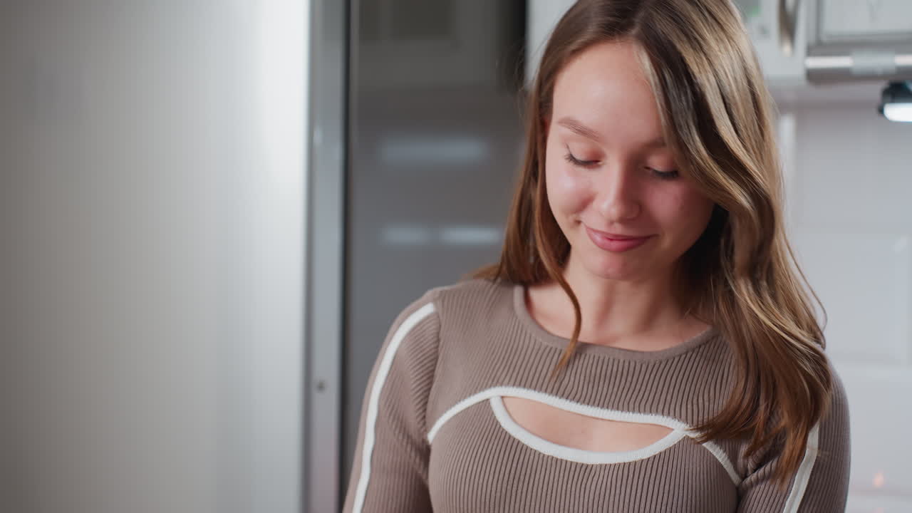 mujer emocionada con cabello castaño rizado en una elegante camiseta marrón baila alegremente en la cocina, el fondo cuenta con una nevera de acero inoxidable y una iluminación cálida