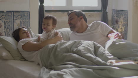 happy parents lying in bed on sunday morning and cuddling their cute baby girl