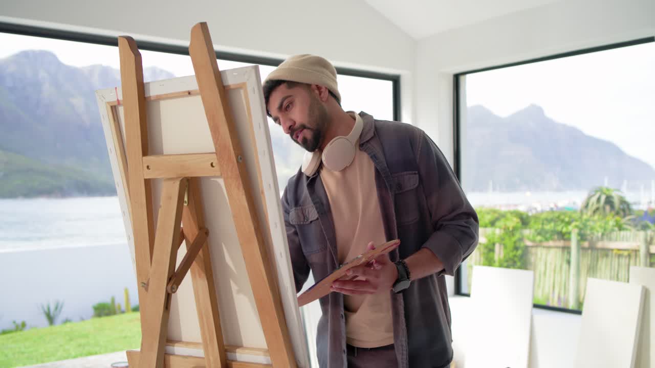 Indian man painting, glancing at lake view for reference, applying strokes at easel with palette