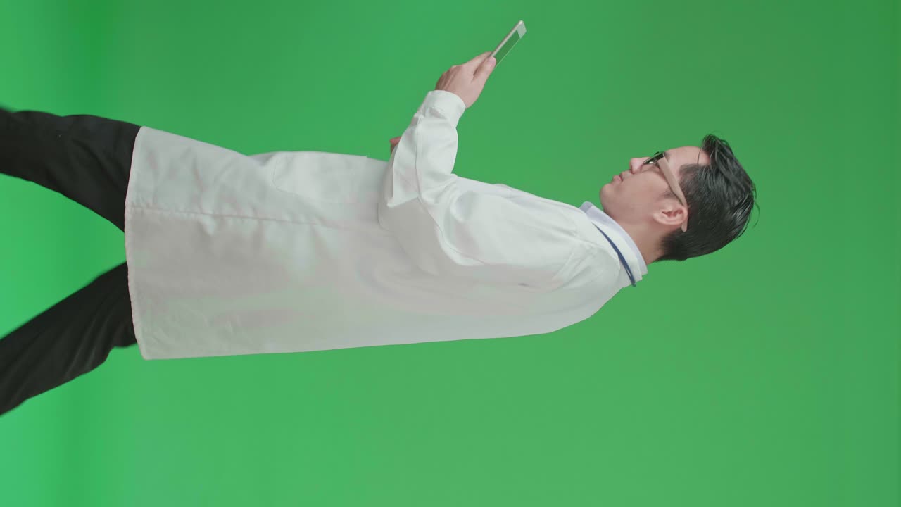 The Side View Of Asian Doctor With Stethoscope Wearing Glasses And White Coat Using Tablet While Walking On Green Screen Chroma Key.