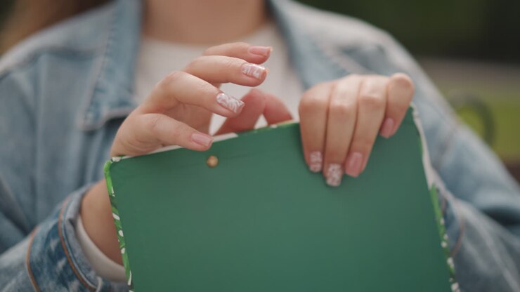 Woman with vibrant nail art. Lady with glittery manicure studying sketches on park bench. Young woman in denim jacket browsing through colorful sketchbook outdoors in peaceful park setting