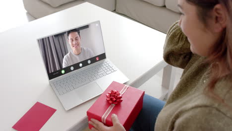 caucasian woman holding red gift using laptop with biracial man on screen