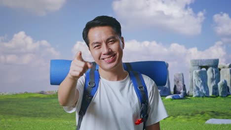 close up of asian male hiker with mountaineering backpack smiling, touching his chest, and pointing to camera while traveling in stonehenge
