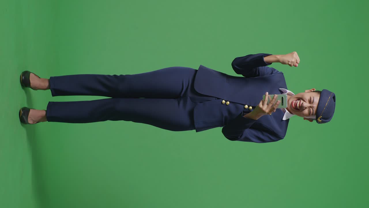 아시아 여성 승무원이 스마트폰을 쳐다보며 소리를 지르며 골을 축하하고 녹색 스크린 배경 스튜디오에 서 있습니다.