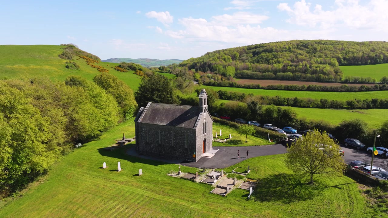 En stenkirke ligger omgivet af frodige marker og bakker i County Laois. Biler er parkeret i nærheden, hvilket indikerer besøgende. Ideel til historiske dokumentarer eller rejseindhold