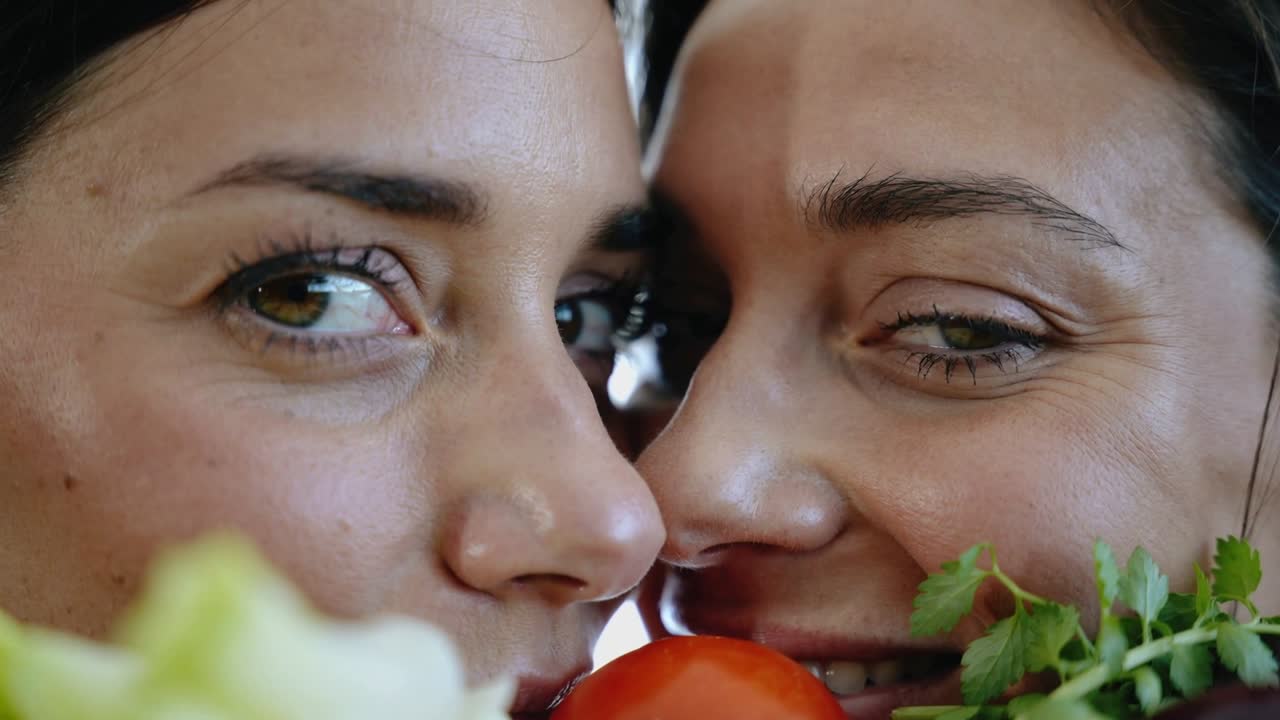 Dos mujeres sosteniendo tomate, lechuga y hierbas, posando mientras la cámara enfoca en la cocina