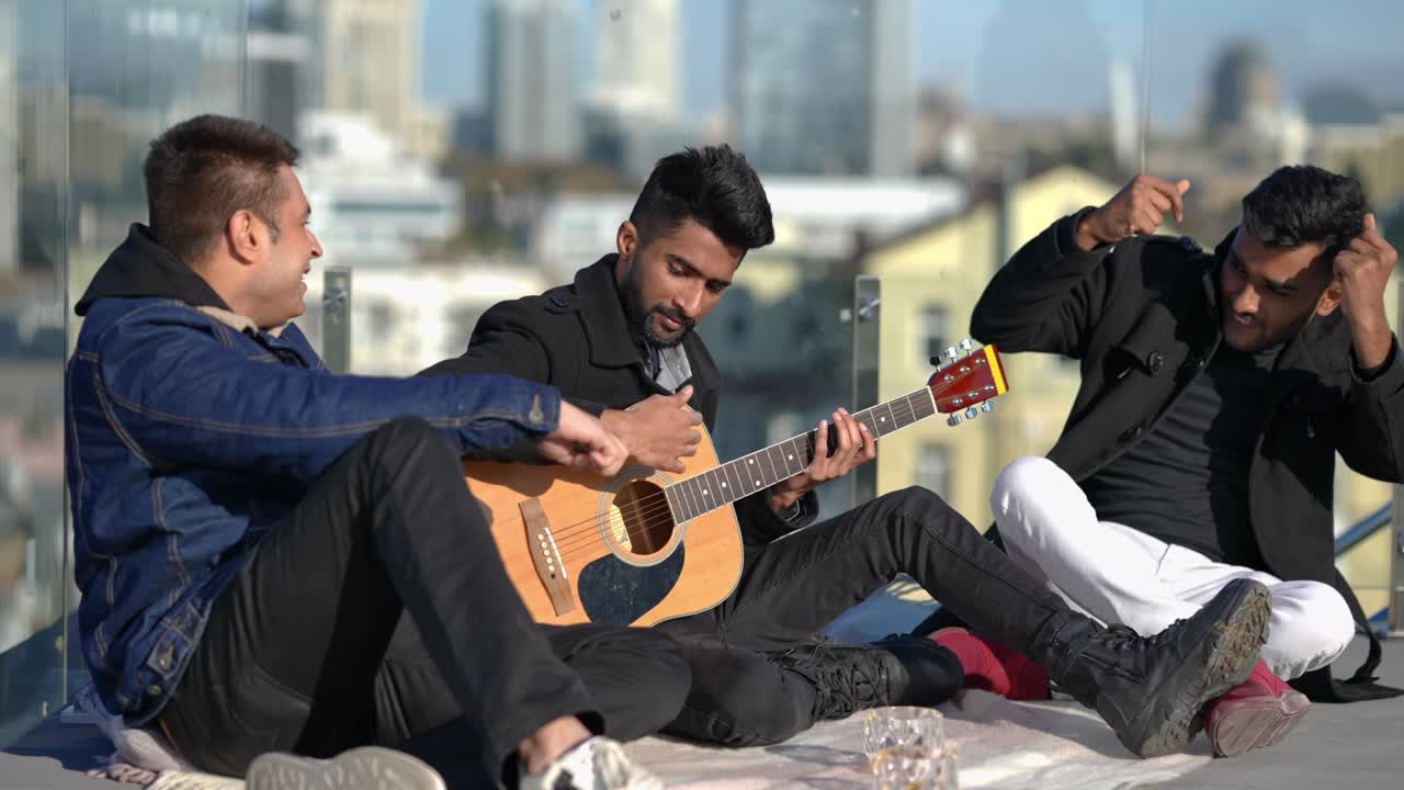 Wide shot confident relaxed man playing guitar as friends dancing sitting on rooftop in sunshine. Happy carefree Middle Eastern men enjoying leisure outdoors on sunny day in urban city.