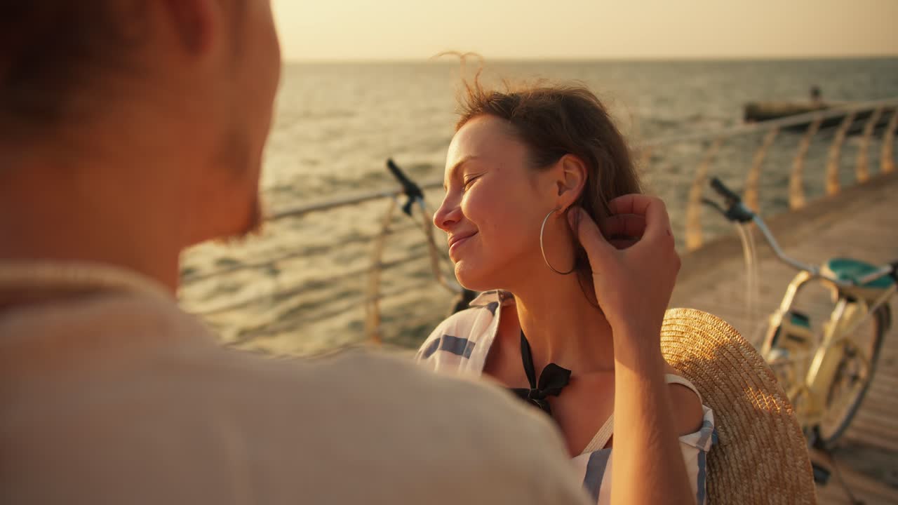 Close-up shot of a guy in a white shirt straightening his hair and near the ear of his girlfriend who smiles and strokes his cheek on the beach near the sea at Sunrise in summer