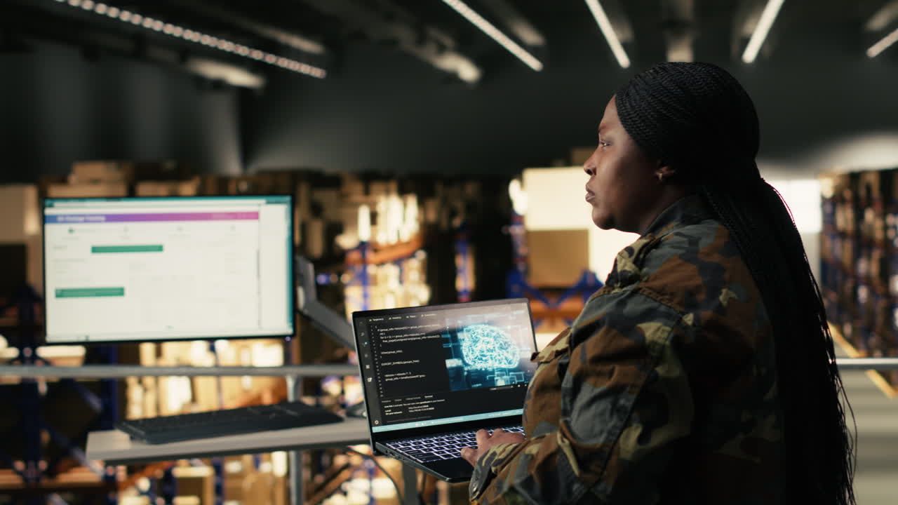 Kvinde i camouflaguniform arbejder på bærbar computer på et lager.