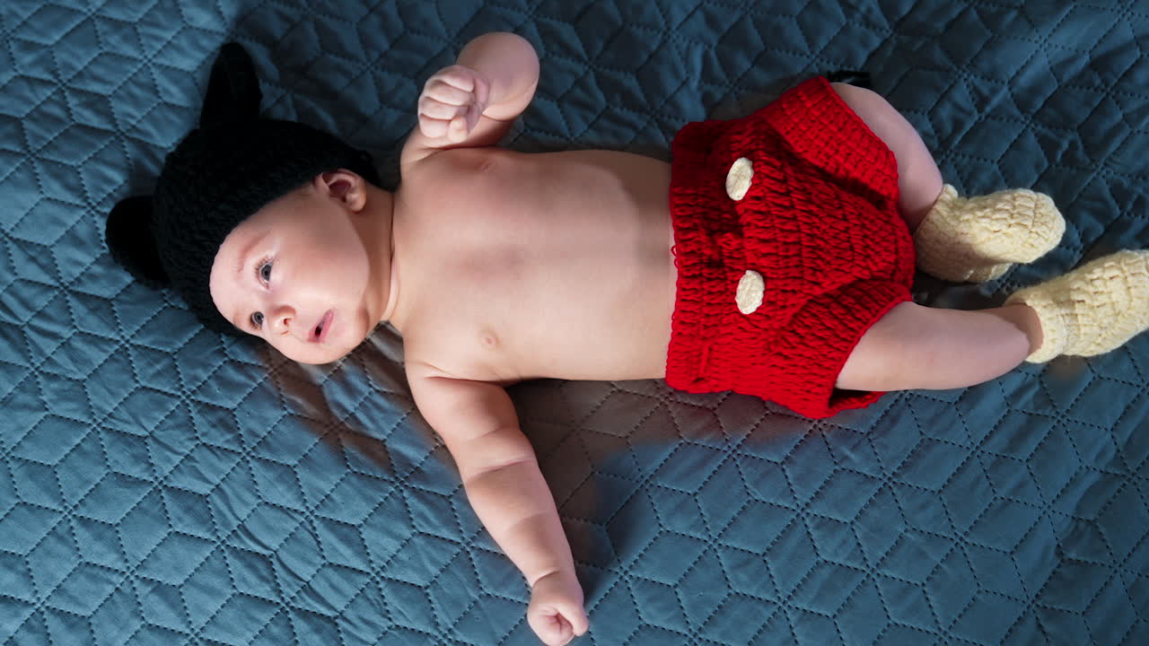 Little toddler wearing funny knitted costume of Mickey Mouse on the bed. Baby boy is unhappy about something and tossing his feet and starting to cry.