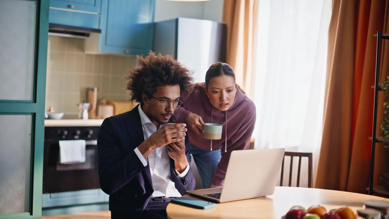 Couple prenant le petit-déjeuner dans la cuisine à la maison. Une femme apporte du café à son petit ami qui travaille sur son ordinateur portable.