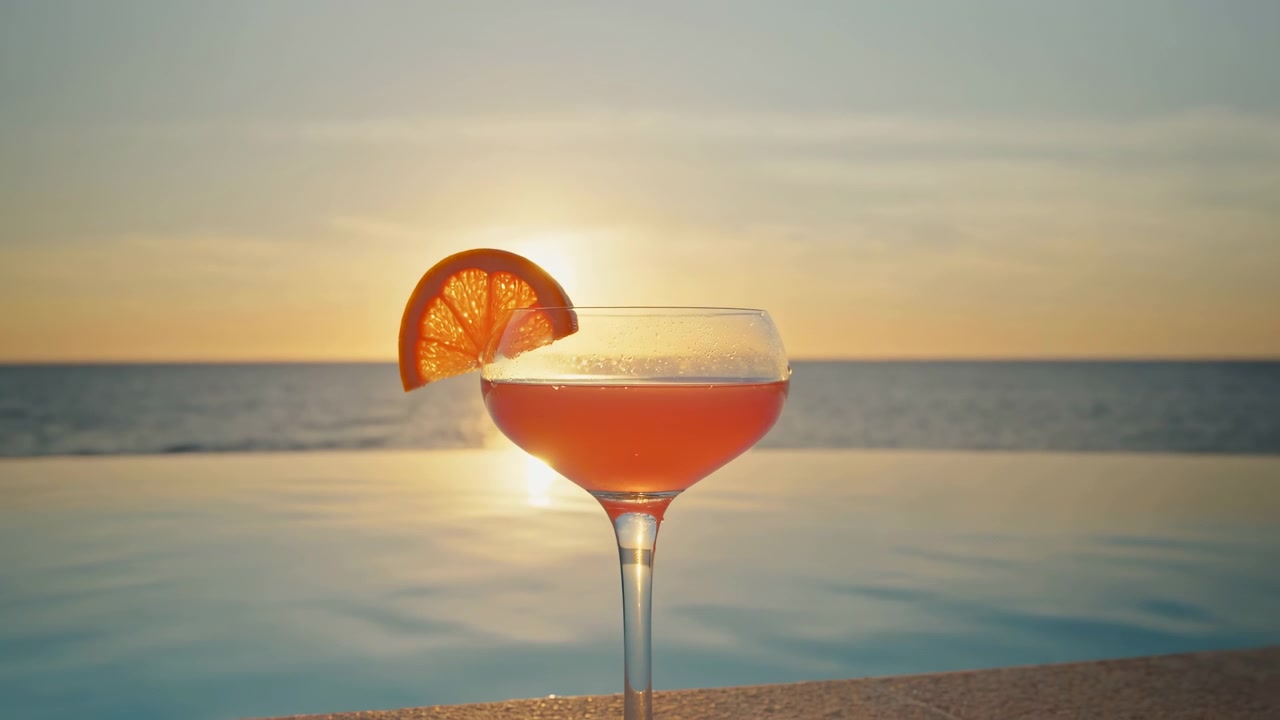 Una vista che si allarga lentamente rivela maggiormente il bordo piscina sotto una bevanda fredda rosso-arancio, mentre una fetta di agrume brilla in controluce e il sole dorato si riflette sull'acqua calma verso l'orizzonte marino