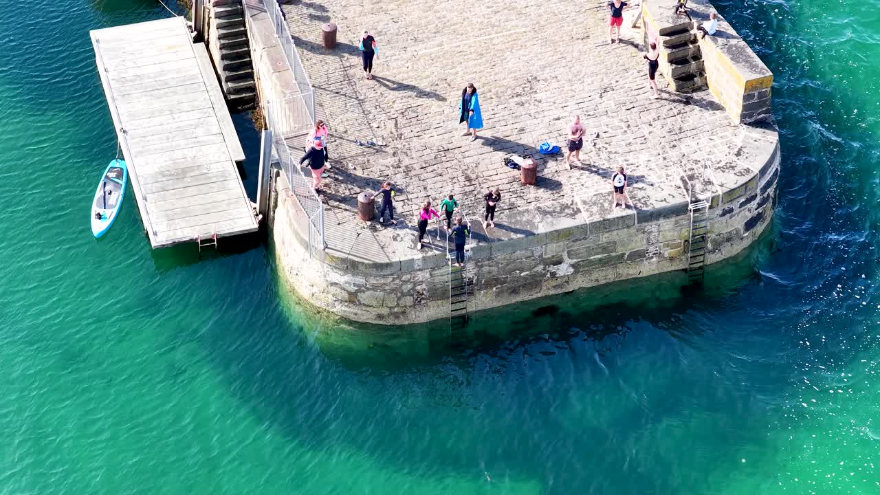 Aerial view of families swimming, gathering, and relaxing on a sunlit stone pier in Scotland