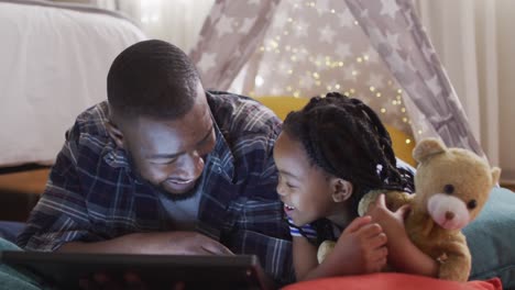 Happy-african-american-father-and-daughter-lying-in-tent-and-using-tablet-with-copy-space