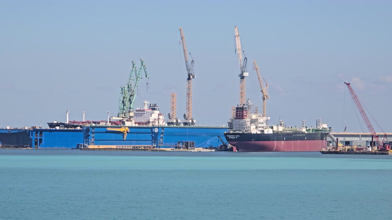 Stor industriell varvsanläggning med en flytande torrdocka och höga kranar. Ett massivt lastfartyg ligger dockat för underhåll och reparation i det ljusblå havsvattnet en solig dag