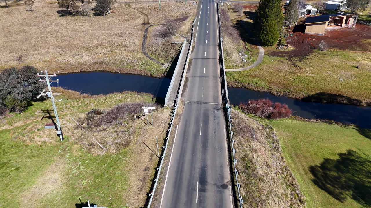 Drone camera smoothly flies above a country road bridge crossing a stream, revealing rural landscape, scattered houses, and clear daylight shadows