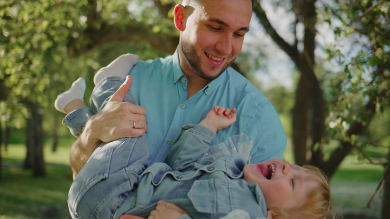 Un uomo tiene in braccio un bambino sorridente in un parco, circondato da alberi verdi. Il padre solleva la figlia, entrambi ridono gioiosamente. La scena cattura un momento familiare giocoso e affettuoso all'aperto. Ideale per pubblicità e contenuti sulla genitorialità.