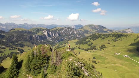 drone circling around a young, strong and fit man standing on the edge overlooking a small, tiny lake below in beautiful turquoise and blue colors with green fields and forest around on a hot day