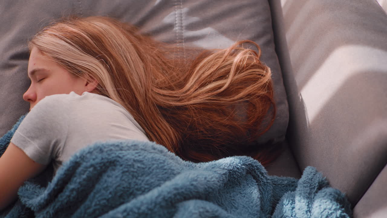 Close up top down view of woman sleeping with blanket under cheek as she dreams peacefully on grey couch while soft daylight illuminates scene and long hair spreads gently behind her