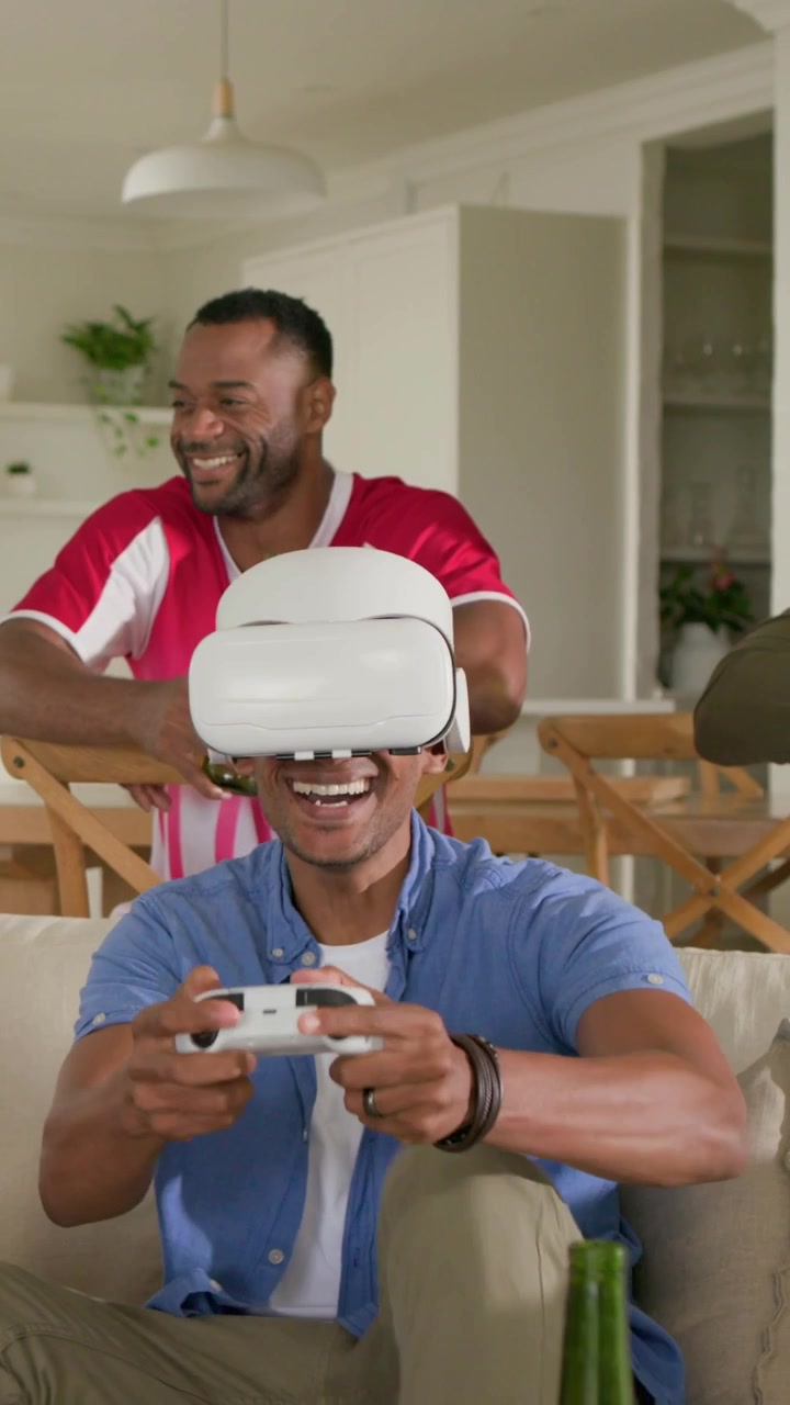 Video verticale: Giocatore in maglietta blu che sistema il visore VR e gioca con un gamepad sul divano, con un amico con una birra.