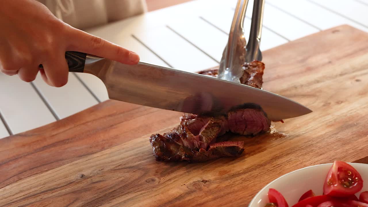 A person slices a cooked beef steak on a wooden board in a bright kitchen setting