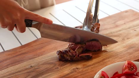 a person slices a cooked beef steak on a wooden board in a bright kitchen setting