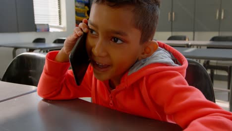 Vista-Frontal-De-Un-Colegial-Asiático-Hablando-Por-Teléfono-Móvil-En-El-Escritorio-Del-Aula-En-La-Escuela-4k