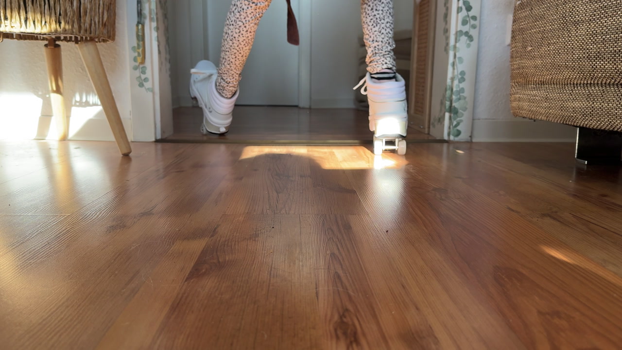 Low-angle view of a child roller skating indoors and falling in a funny, playful moment. Captures humor, movement, and candid childhood activity on a wooden floor in warm home lighting.