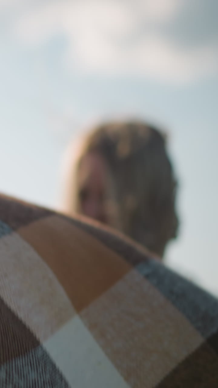 una mujer con el rostro ligeramente borroso y el cabello cubriendo su cara sostiene una bufanda a cuadros mientras el viento la sopla suavemente. la escena captura un momento sereno de conexión con la naturaleza