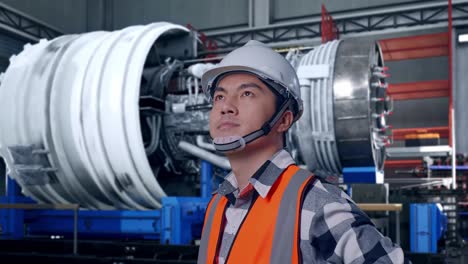 close up side view of asian male engineer wearing safety helmet looking around while standing with arms akimbo with airplane engine maintenance conducted, analytics checking the turbine