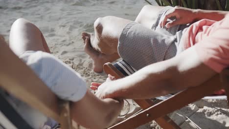 Happy-diverse-couple-sitting-in-deckchairs-holding-hands-on-beach-in-the-sun,-in-slow-motion