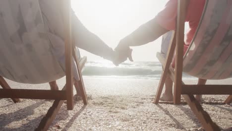 back view of hispanic senior couple relaxing on sunbed on beach at sunset, holding hands