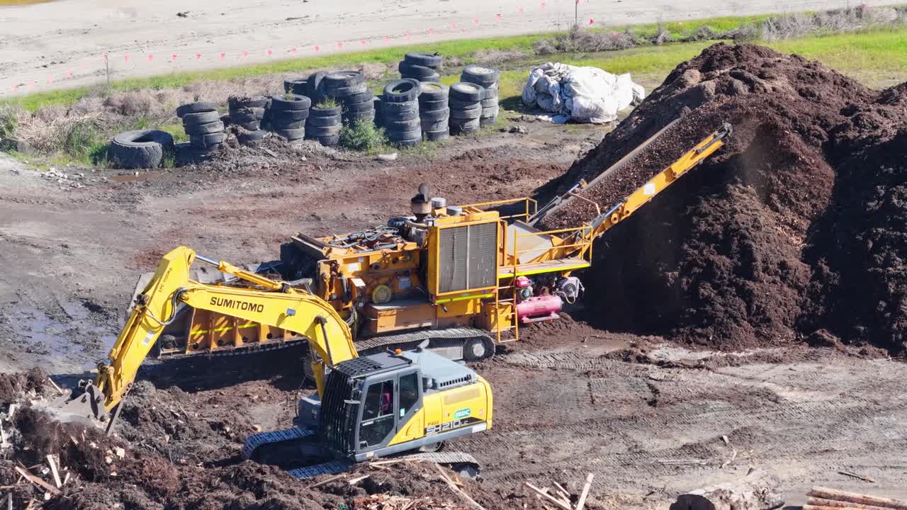 Drone footage captures an excavator loading mulch into a processing machine in a sunny, industrial setting