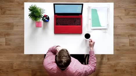 young man in glasses looking on laptop's screen and drinking coffee, topshot, sitting behind desk